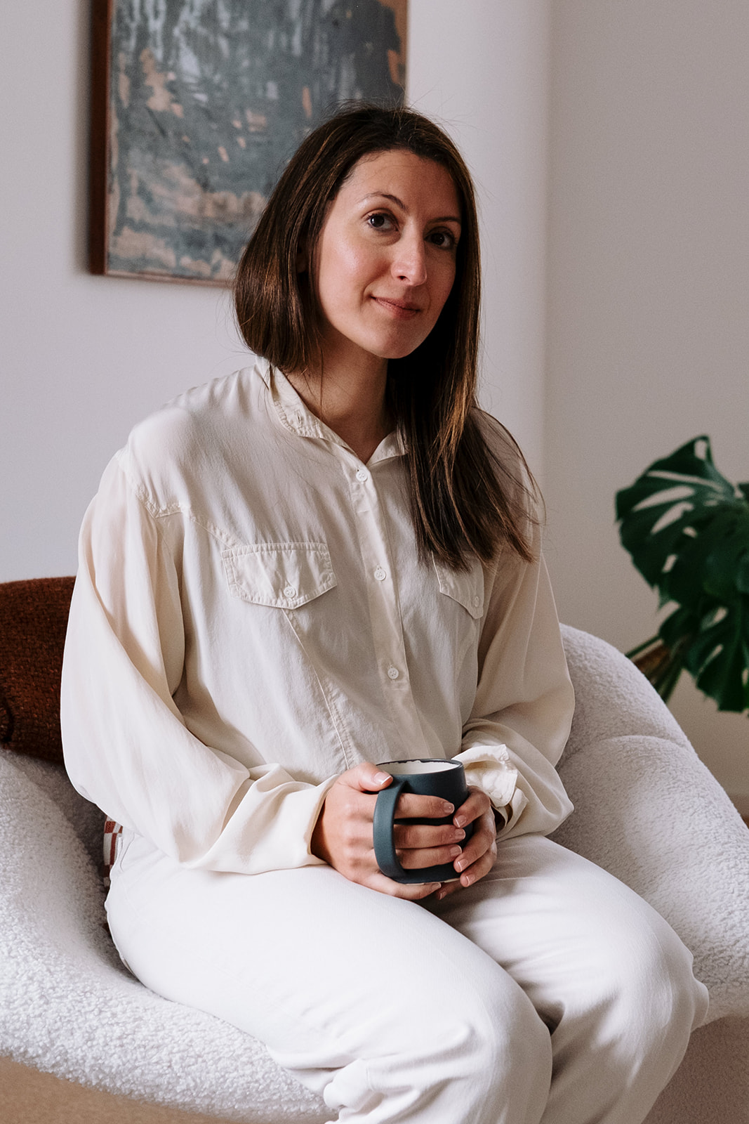 Sarah Miloudi, seated in a white armchair and holding a mug in both hands, looking at the camera with a slight smile.