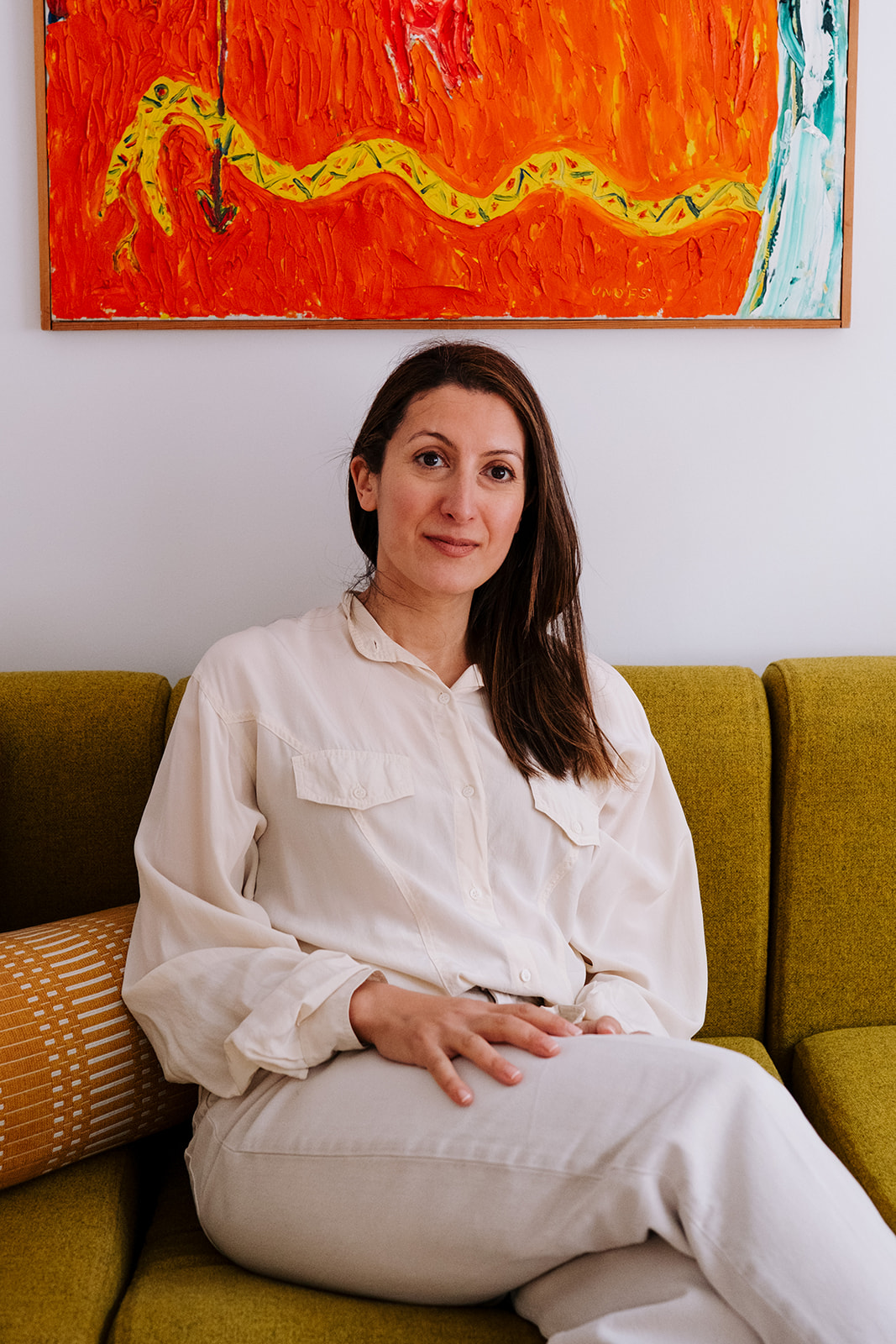 Sitting on a green sofa beneath a bright orange painting, one hand resting on her knee, looking at the camera with a slight smile.